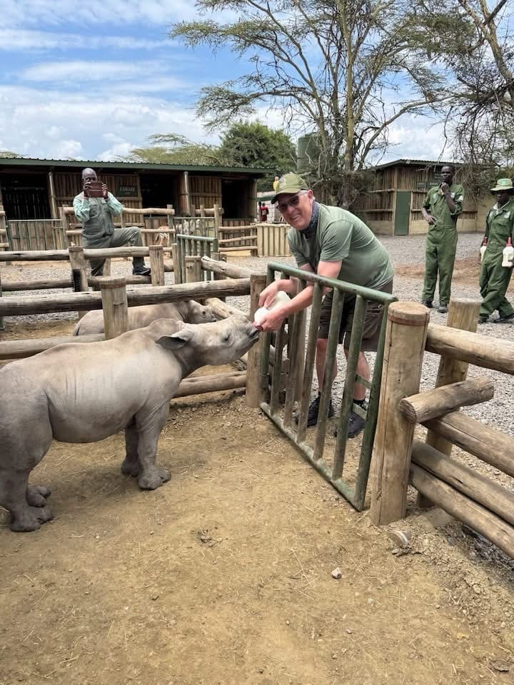 P1040783 Man in a green shirt feeds a young rhinoceros through a wooden enclosure fence at a wildlife sanctuary.