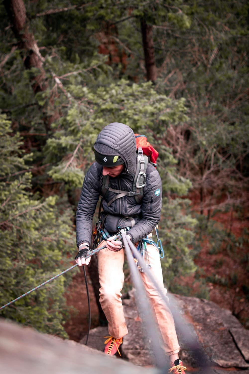 P1040783 Rock climber rappels down a forested cliff wearing a helmet, harness, and backpack.